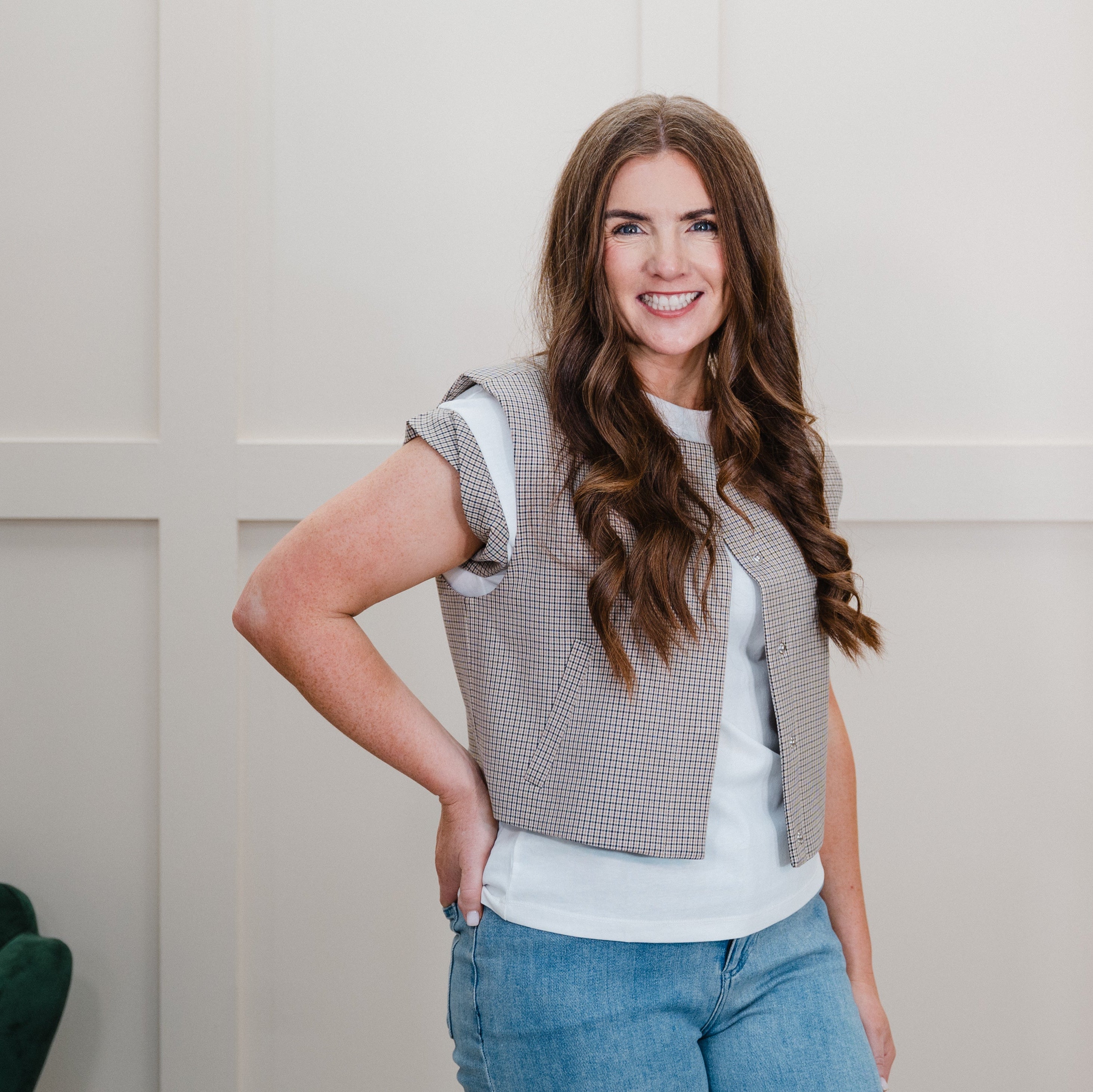 A woman with long brown hair stands indoors wearing a sleeveless gray and white houndstooth vest over a white t-shirt and blue jeans. She is posing in front of a white paneled wall with a gold mirror frame visible on the right.