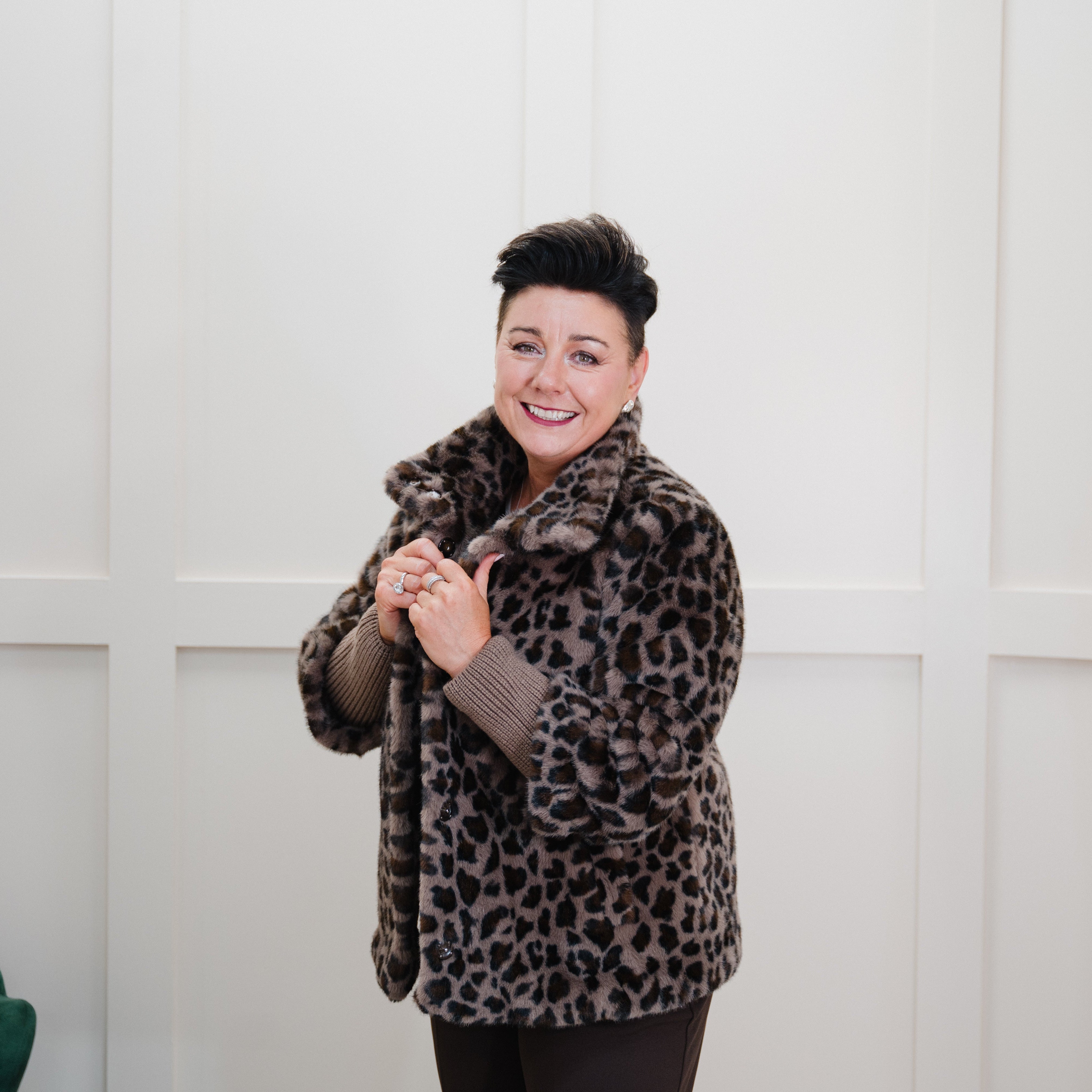 A woman with short dark hair smiling while wearing a brown and black leopard-print faux fur coat in a white paneled room with a gold mirror frame.